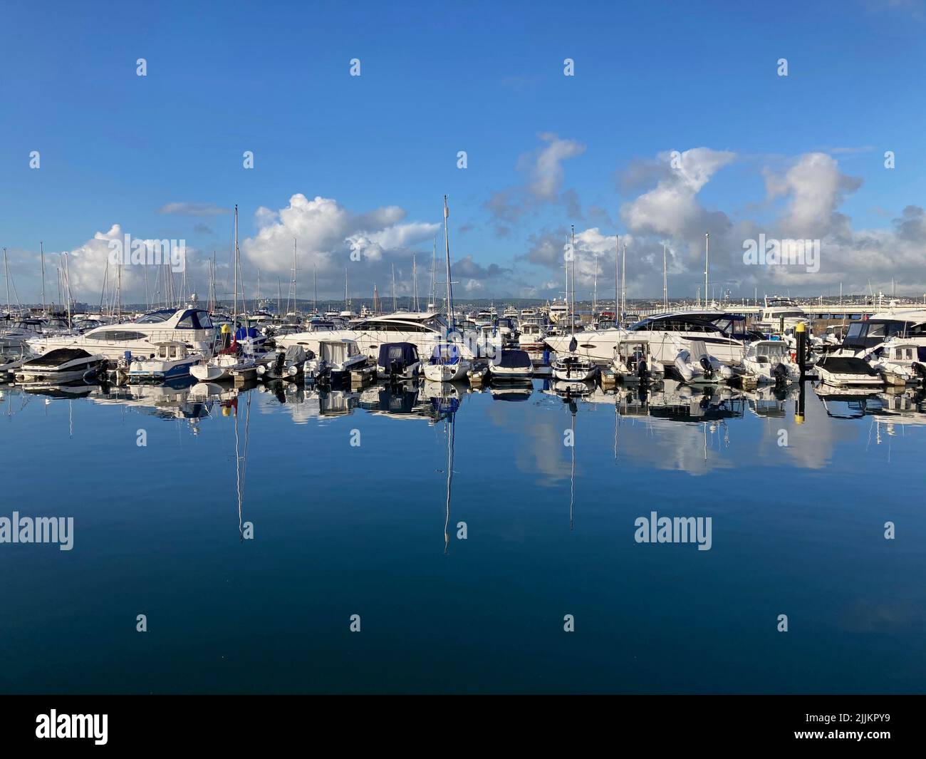 Segelboote und Motoryachten an einem sonnigen Tag in einer Marina. Boote und Masten spiegelten sich im stillen Wasser in einer ruhigen Szene wider. Flauschige Wolken am Himmel. Stockfoto