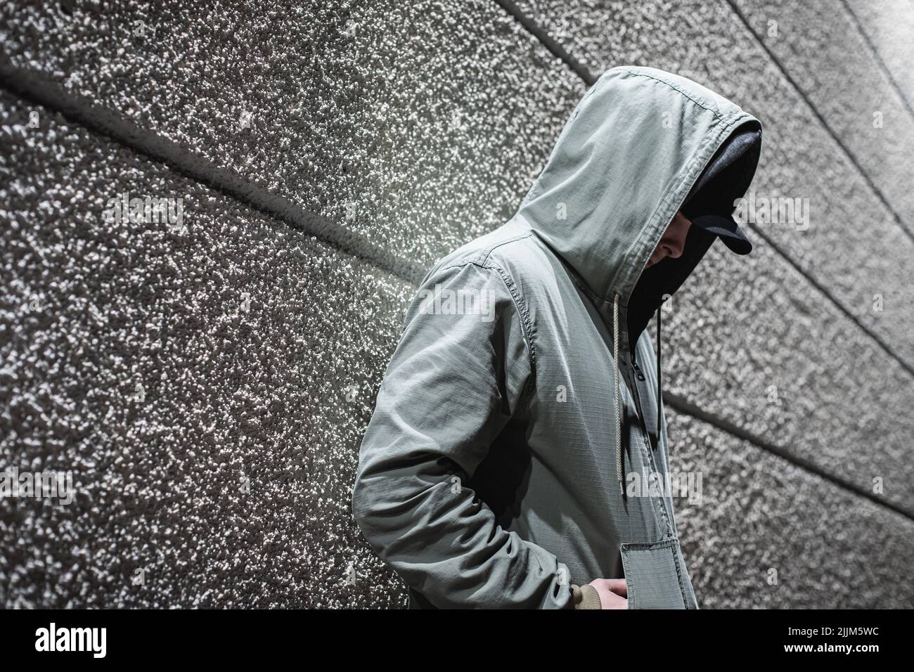 Eine Seitenansicht einer Person in einem Anorak mit den Händen in den Taschen, die an einer gestreiften Wand gelehnt sind Stockfoto