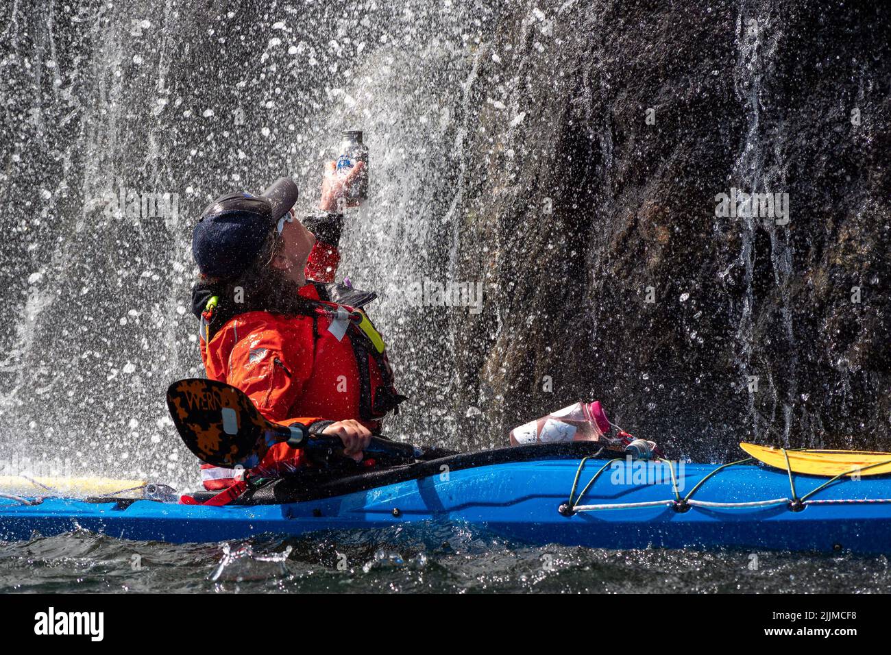 Ein Mädchen füllt ihre Wasserflasche unter einem kleinen Wasserfall, während es in ihrem Kajak sitzt Stockfoto