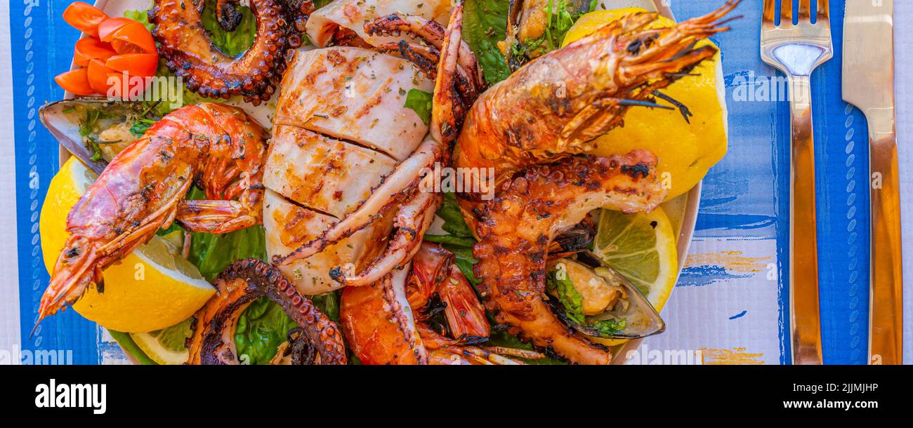 Meeresfrüchteplatte mit gegrilltem Tintenfisch, Krake, Garnelen und Muscheln auf grünem Salat mit Zitrone und Tomaten, Panorama Stockfoto