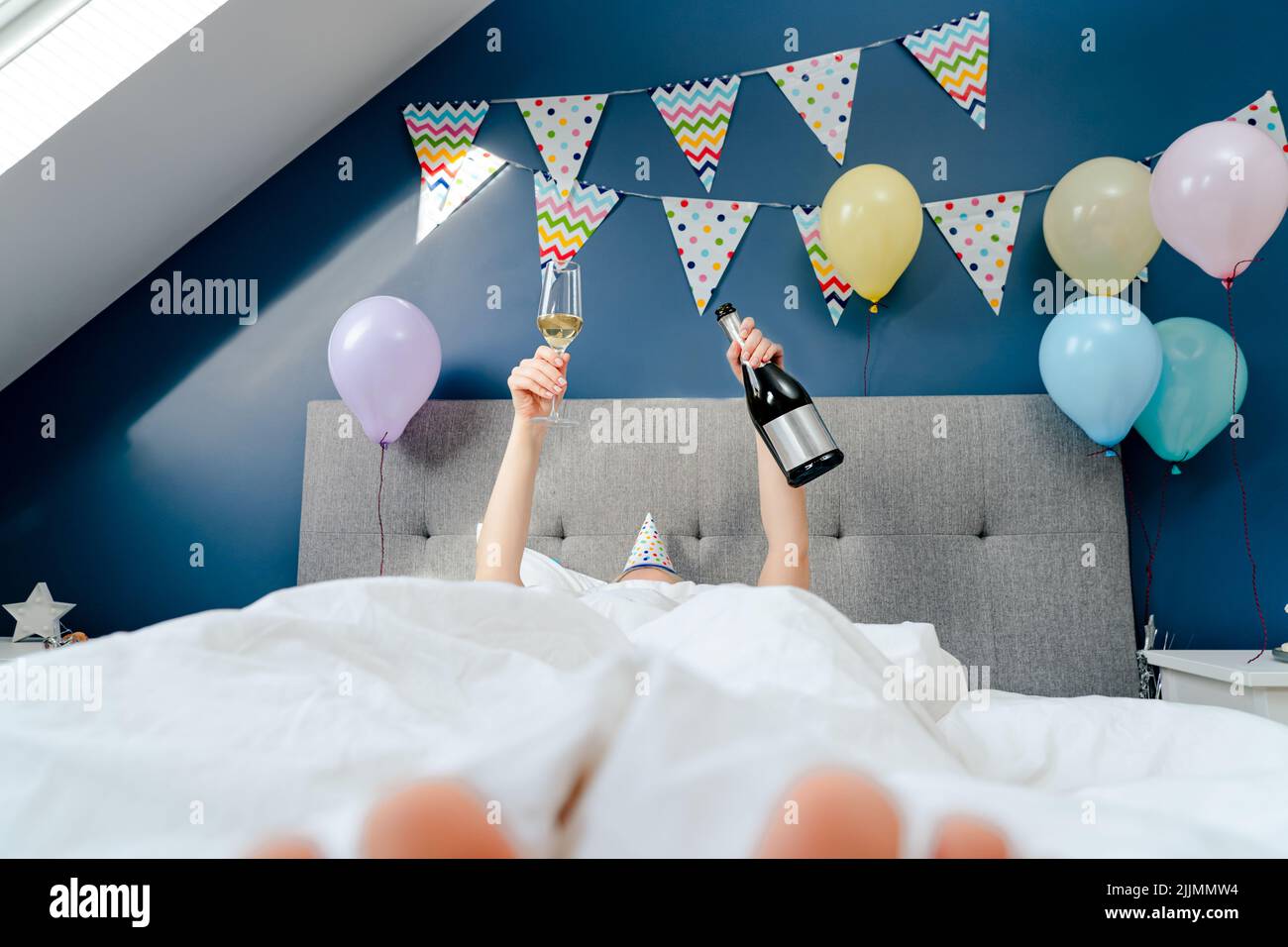 Hände einer Frau mit einer Flasche und einem Glas Champagner liegen auf dem Bett im Schlafzimmer mit Ballons und Girlande geschmückt.Zeit für sich selbst celebratio Stockfoto