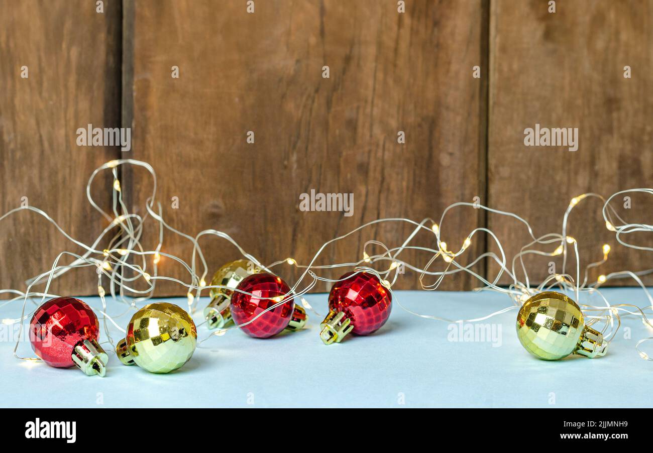 Ein Schuss goldener und roter Weihnachtskugeln mit einer Lichterkette auf einem weißen Tisch an der braunen Holzwand Stockfoto