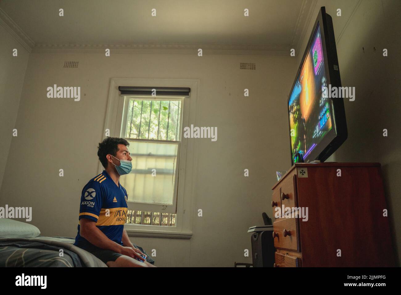 Ein hispanischer Mann, der mit einer blauen medizinischen Gesichtsmaske auf einem Bett sitzt, spielt während der Covid-Isolation in Melbourne, Australien, PlayStation Stockfoto