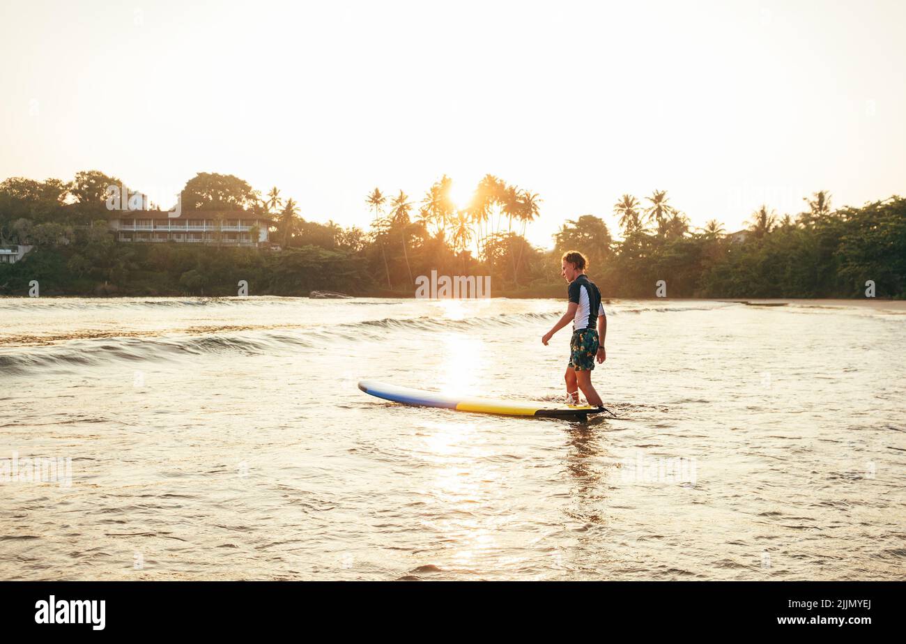 Junger Teenager Junge mit buntem Surfbrett geht ins Meer zum Surfen. Er genießt ein wunderschönes Sonnenuntergangslicht am Dewata Strand auf Sri Lanka. Aktiver Sport Stockfoto