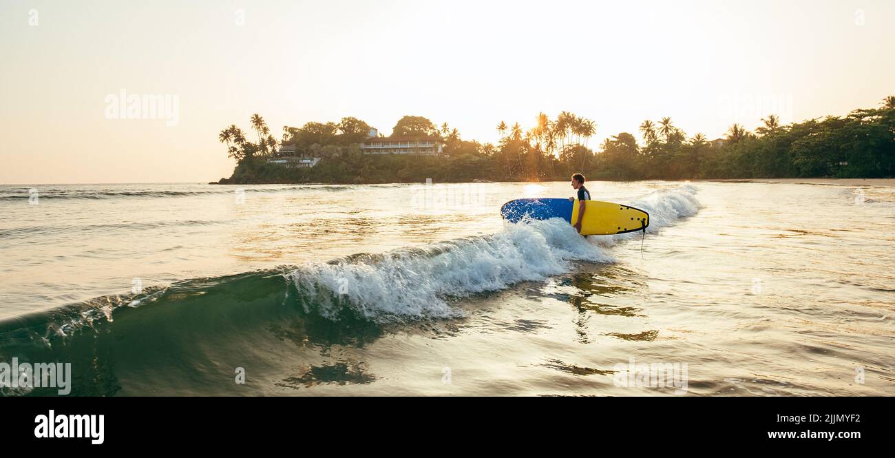 Junger Teenager Junge mit buntem Surfbrett gehen zum schaumig Meer zum Surfen. Er genießt ein wunderschönes Sonnenuntergangslicht am Dewata Strand auf Sri Lanka. Aktiver spor Stockfoto