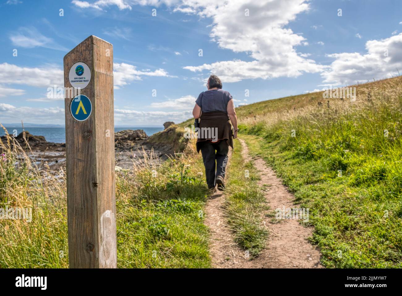 Ein Wegzeichen und Richtungspfeil mit einer Frau, die auf dem Fife Coastal Path in St. Monans im Osten von Fife, Schottland, läuft. Stockfoto