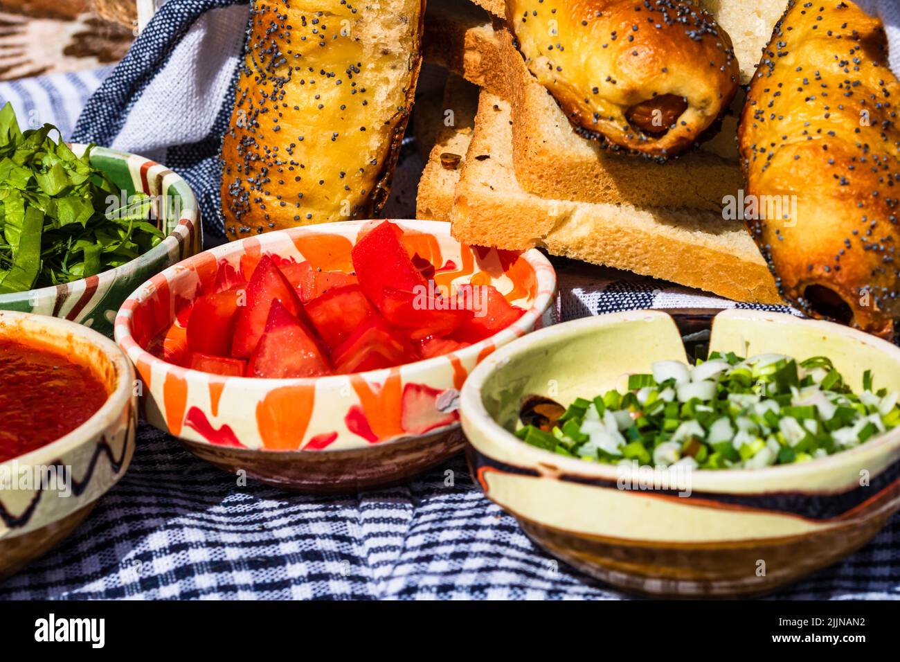 Rustikale Zusammensetzung mit Würstchenbrötchen und verschiedenen Schüsseln mit Sauce und gehacktem Gemüse (Tomaten, grüner Salat, grüne Zwiebel, grüner Knoblauch) Stockfoto