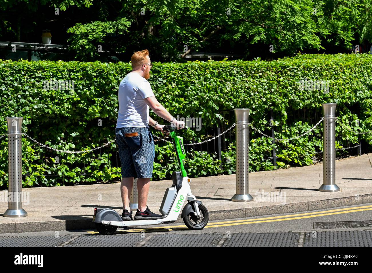 London, Großbritannien - 2022. Juni: Person auf einem Elektroroller, die auf einer Straße entlang einer grünen Hecke um Gärten in Canary Wharf fährt Stockfoto