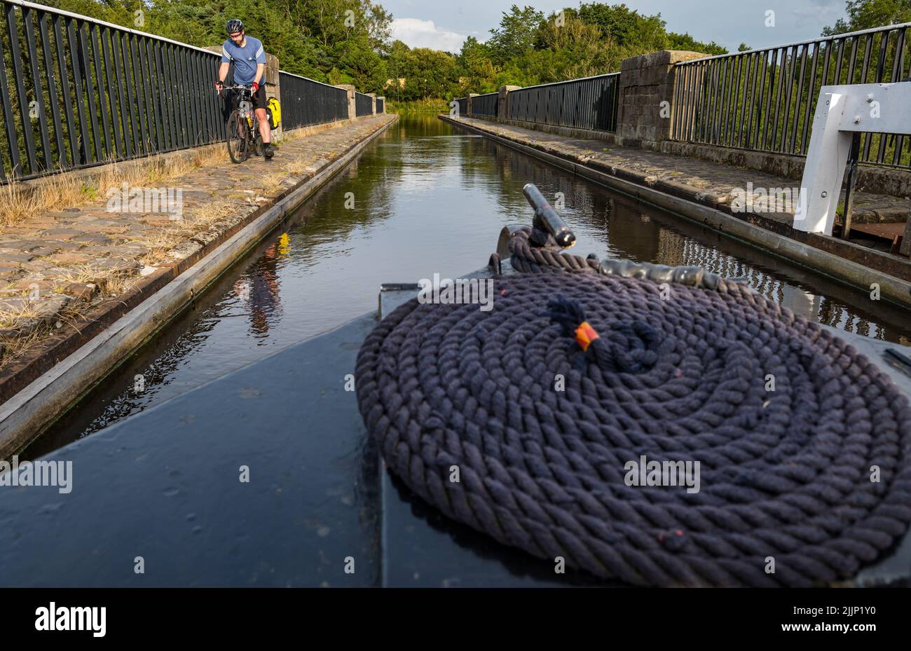 Schottland, Vereinigtes Königreich, 27.. Juli 2022. UK Wetter: Sonniger Abend auf dem Union Canal. Ein Radfahrer, der auf dem Kanalpfad Fahrrad fährt, während ein enges Boot das Almond Aqueduct in West Lothian überquert Stockfoto