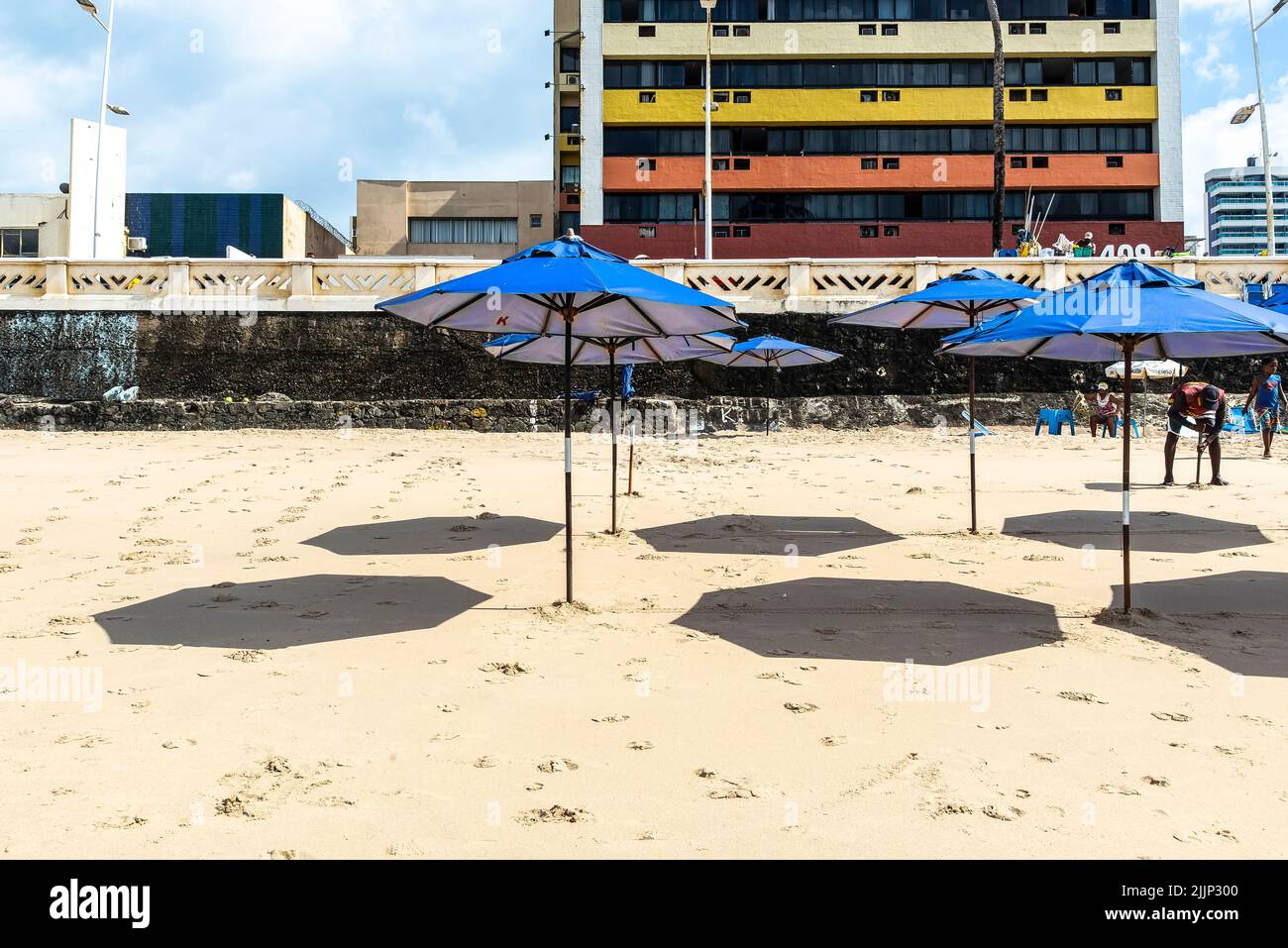 Eine Reihe von blauen Sonnenschirmen mit Schatten auf Sand am Strand Farol da Barra in Salvador, Bahia, Brasilien. Stockfoto