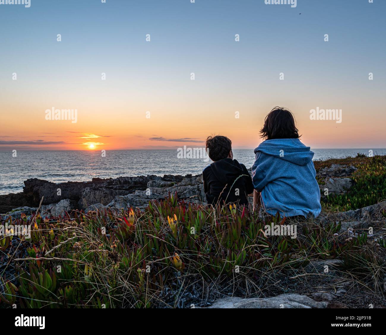 Zwei Kinder sitzen auf den Felsen und beobachten den Sonnenuntergang auf Cascais, Lissabon Stockfoto