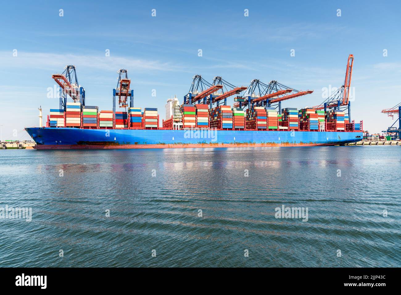 An einem sonnigen Sommertag wird das Containerschiff verladen, während es an einem Containerdock mit hohen Portalkranen befestigt wird. Stockfoto