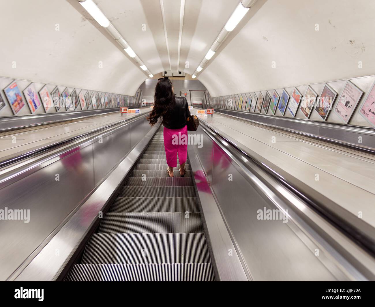 London, Greater London, England, Juli 02 2022: Dame mit rosa Hosen und schwarzer Jacke auf einer Rolltreppe an einer ansonsten leeren U-Bahnstation Stockfoto