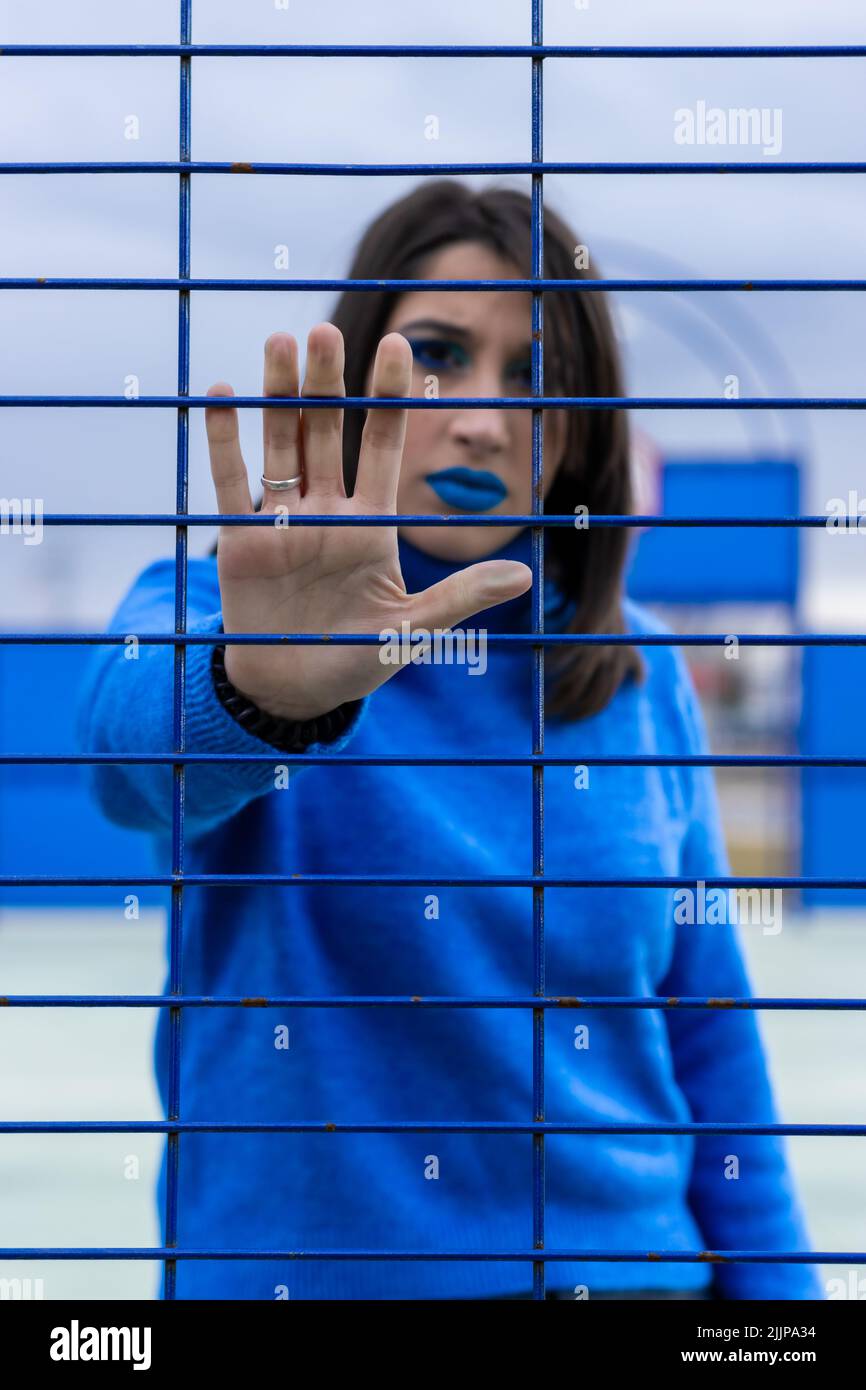 Ein vertikales Porträt einer ernsthaften kaukasischen Frau in blauer Kleidung hinter einem Zaun mit Stoppschild-Geste mit ihrer Hand. Stockfoto