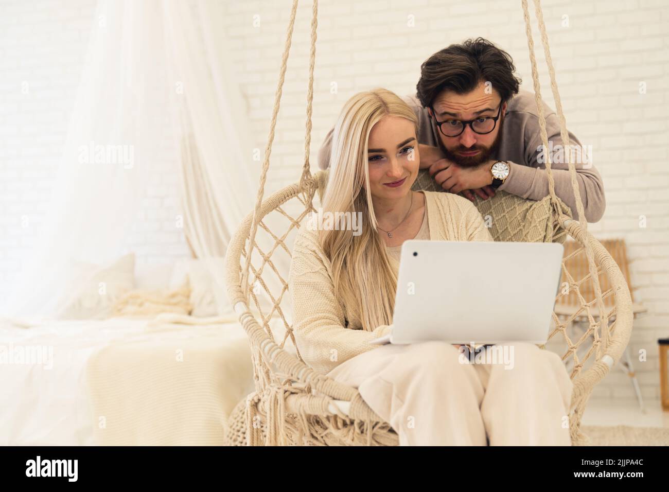 Kaukasisches Paar im Schlafzimmer. Dunkelhaariger Mann mit Bart und Brille, der seine blonde Freundin mit einem Laptop-Computer im Hängesessel beobachtet. Innenaufnahme. Hochwertige Fotos Stockfoto