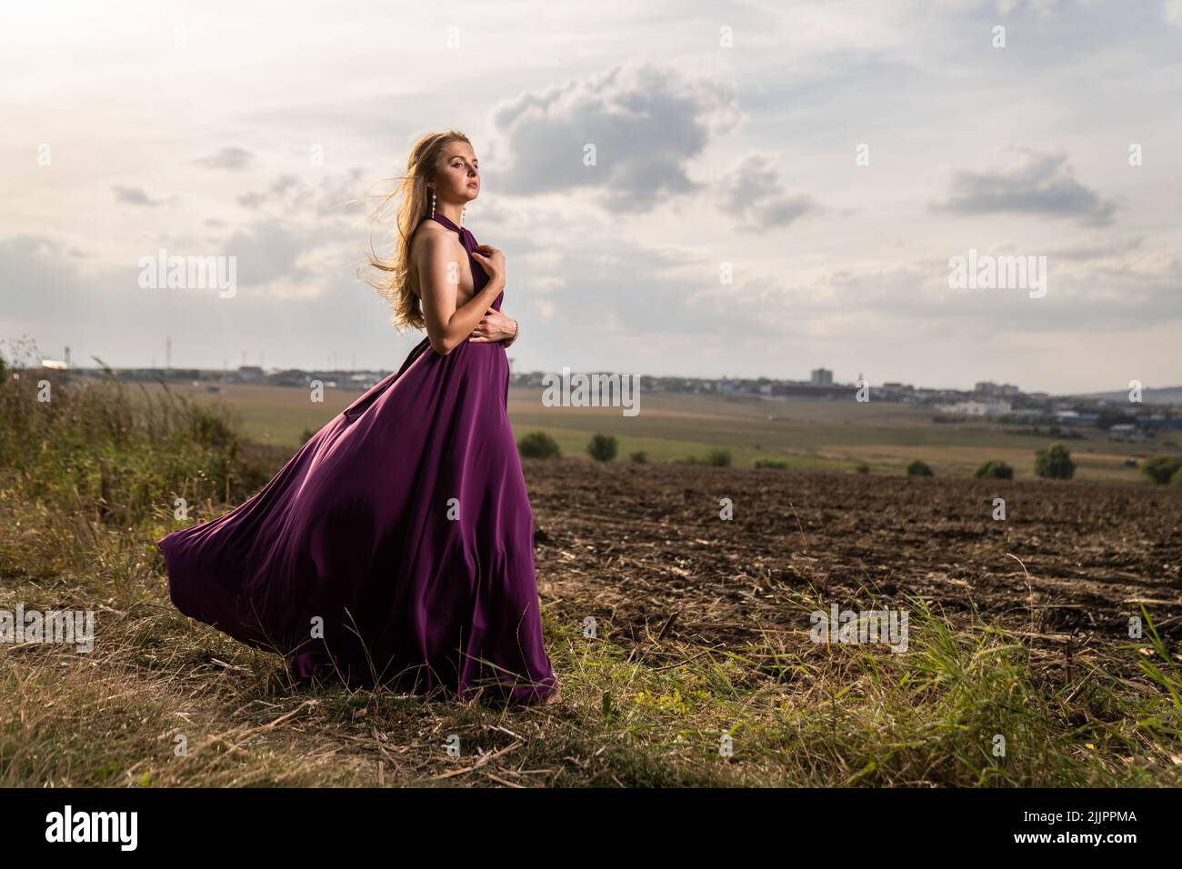 Eine schöne Aufnahme einer kaukasischen Frau in einem eleganten violetten Kleid, das auf einem Feld unter den Wolken posiert Stockfoto