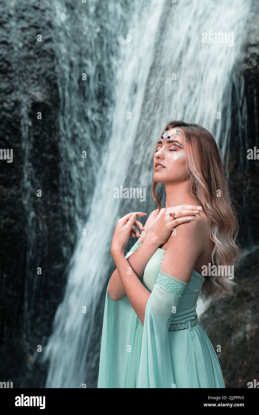 Eine vertikale Aufnahme eines jungen Weibchens in einem grünen Kleid und Stirnschmuck, der sich in der Nähe eines Wasserfalls posiert Stockfoto