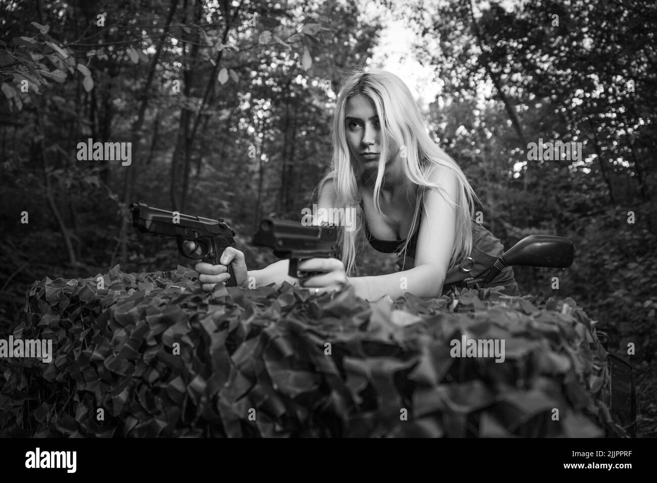 Ein Porträt einer kaukasischen blonden Frau, die auf einem Fahrrad sitzt und in Graustufen in einem Wald Waffen hält Stockfoto