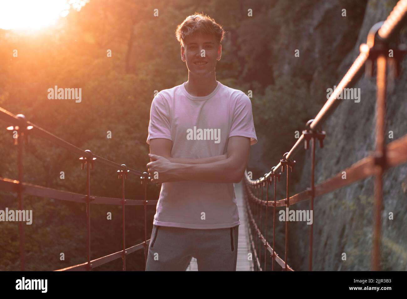 Ein flacher Fokus eines fröhlichen jungen, hübschen kaukasischen Mannes, der auf einer Brücke gegen die Sonnenstrahlen posiert Stockfoto