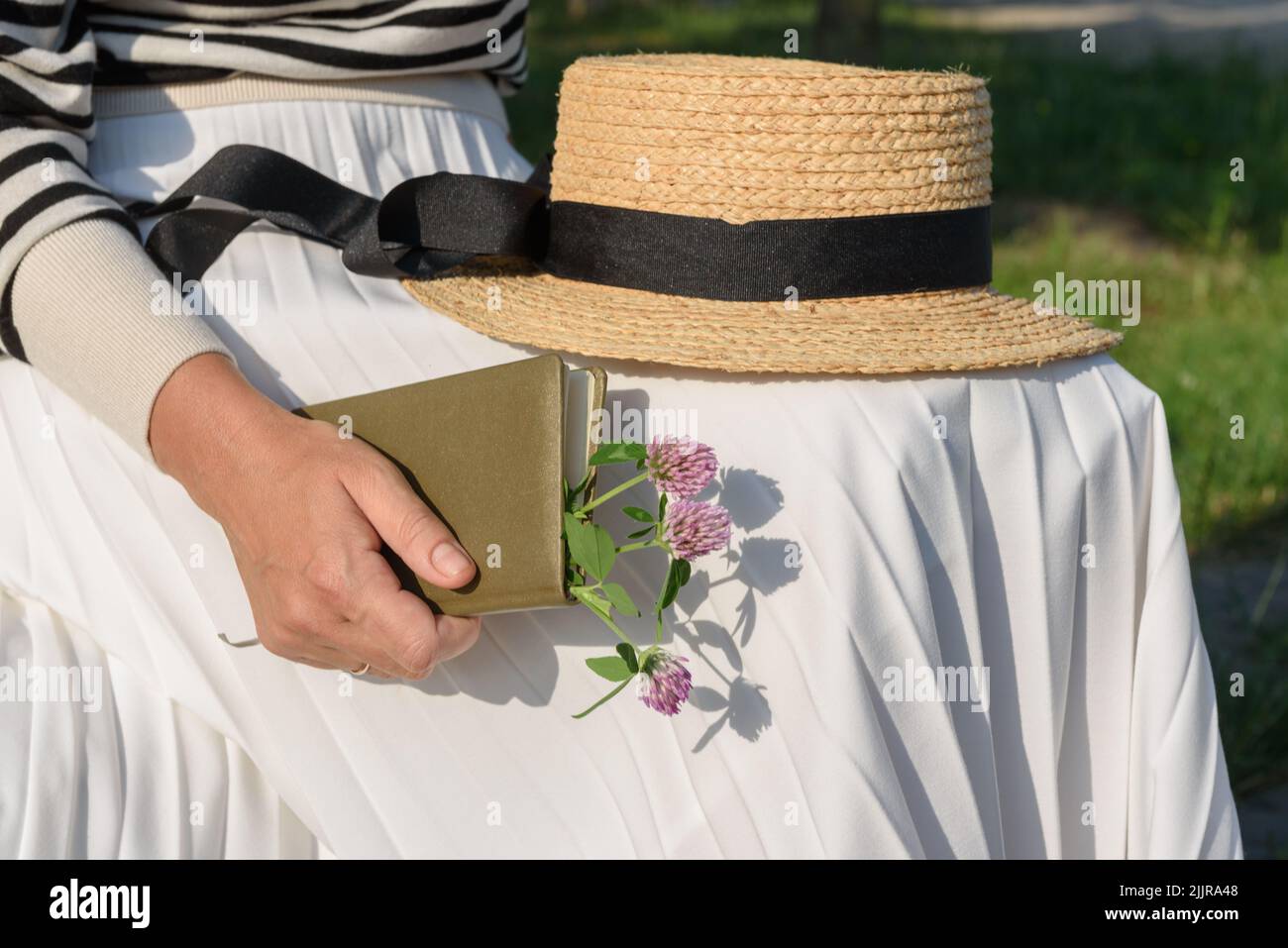 Verheiratete Frau, die in einem Park mit einem Bootsfahrer auf dem Schoß sitzt, mit geschlossenem olivfarbenem Tagebuch mit Blumenstrauß aus Kleeblatt im Inneren, beschnittene Ansicht. Stockfoto