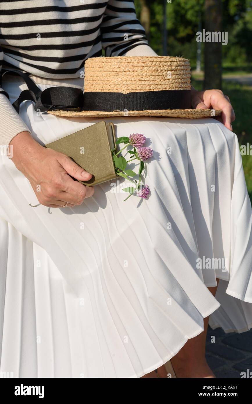 Verkürzte Ansicht der Frau in gestreiftem Pullover und weißem Faltenrock, die im Park mit Bootsfahrer auf dem Schoß sitzt und Tagebuch mit Blumenstrauß im Inneren hält Stockfoto