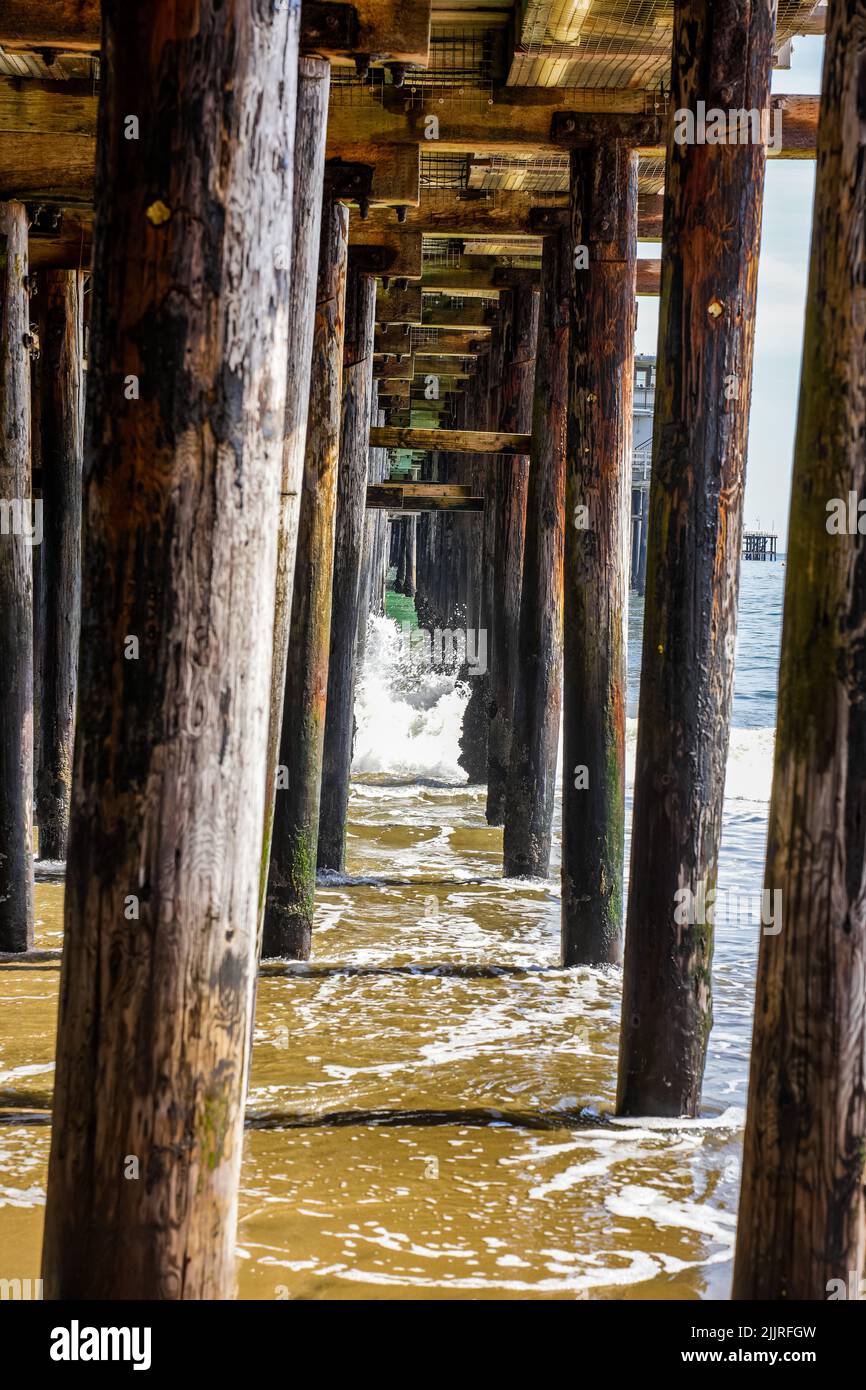 Eine vertikale Aufnahme von Holzsäulen eines Decks mit Wellen, die die sandige Küste waschen Stockfoto