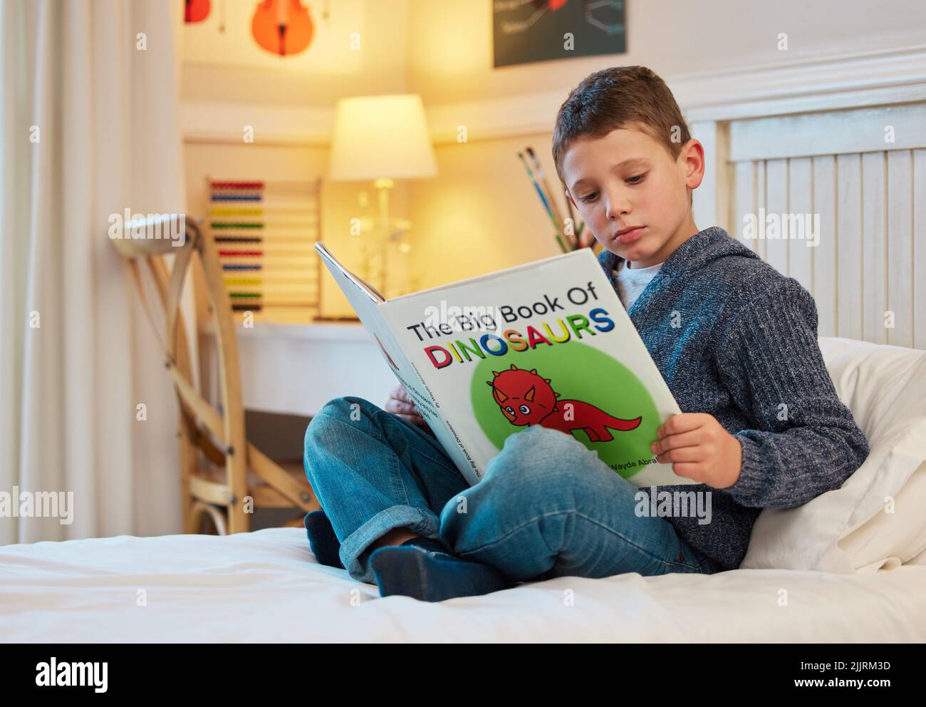 Er hat ein intensives Interesse an Dinosauriern. Ein kleiner Junge liest ein Buch über Dinosaurier in einem Schlafzimmer zu Hause. Stockfoto