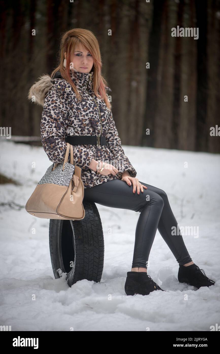 Eine kaukasische junge Frau, die einen Wintermantel mit Leopardenmuster trägt und auf einem Autoreifen in einem verschneiten Park sitzt Stockfoto