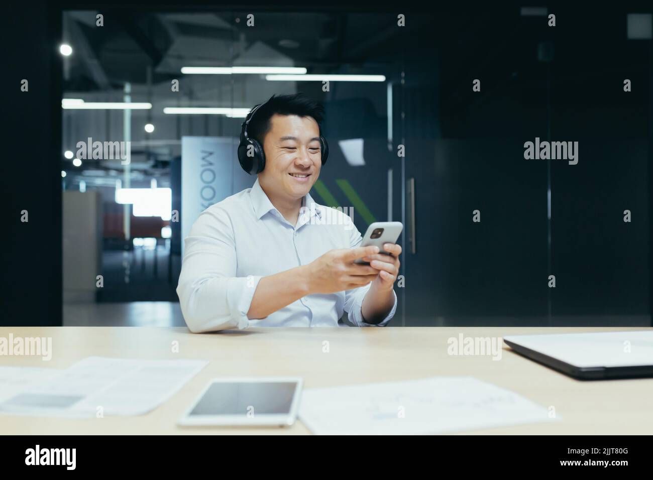 Eine Pause bei der Arbeit. Junger gutaussehender Asiatischer, Freiberufler, der mit Kopfhörern Musik am Telefon hört. Tanzt mit, lächelt. Sitzen an einem Schreibtisch in einem modernen Büro Stockfoto