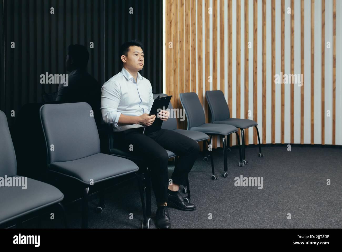 Ein junger, gutaussehender asiatischer Mann wartet in der Empfangshalle auf ein Interview für einen Job in einem Büro, einer Bank oder einem Unternehmen. Er hält eine Mappe in den Händen, sorgt sich, schüttelt sie ab Stockfoto