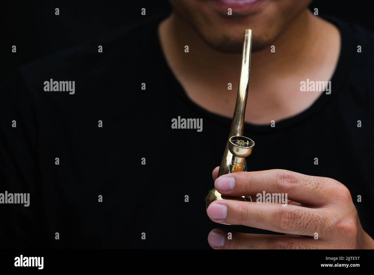 Ein asiatischer Mann raucht zu Hause aus einer Pfeife Marihuana. Studio-Shooting mit Model, das den Rauchtopf mit einer Pfeife vor dunklem Hintergrund simuliert. Legalisierung von Cannabis Stockfoto