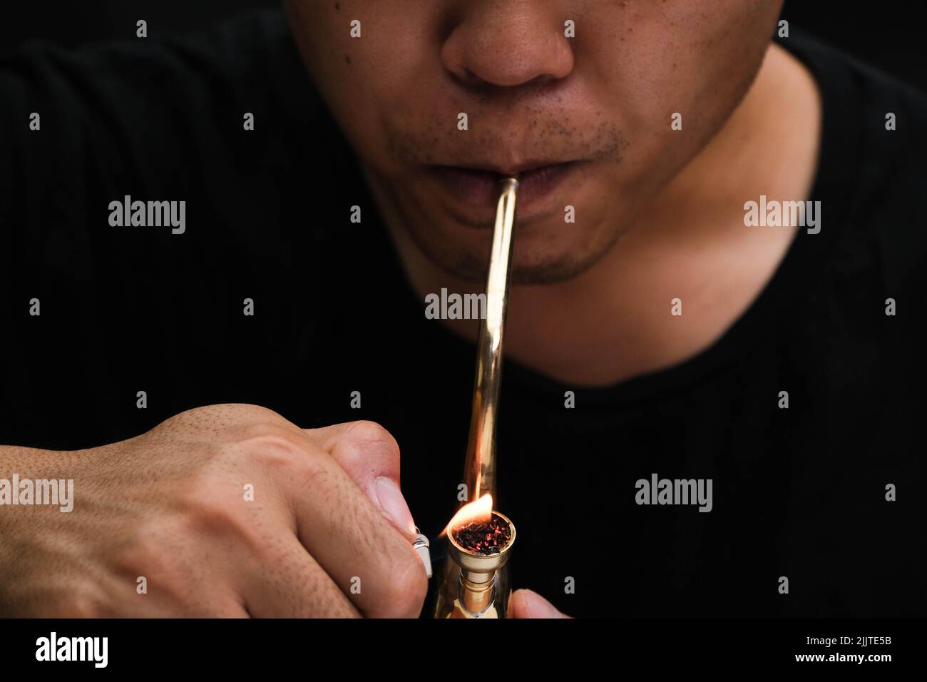 Ein asiatischer Mann raucht zu Hause aus einer Pfeife Marihuana. Studio-Shooting mit Model, das den Rauchtopf mit einer Pfeife vor dunklem Hintergrund simuliert. Legalisierung von Cannabis Stockfoto
