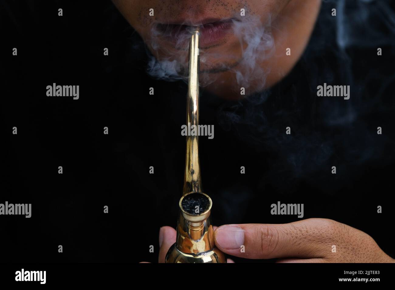 Ein asiatischer Mann raucht zu Hause aus einer Pfeife Marihuana. Studio-Shooting mit Model, das den Rauchtopf mit einer Pfeife vor dunklem Hintergrund simuliert. Legalisierung von Cannabis Stockfoto