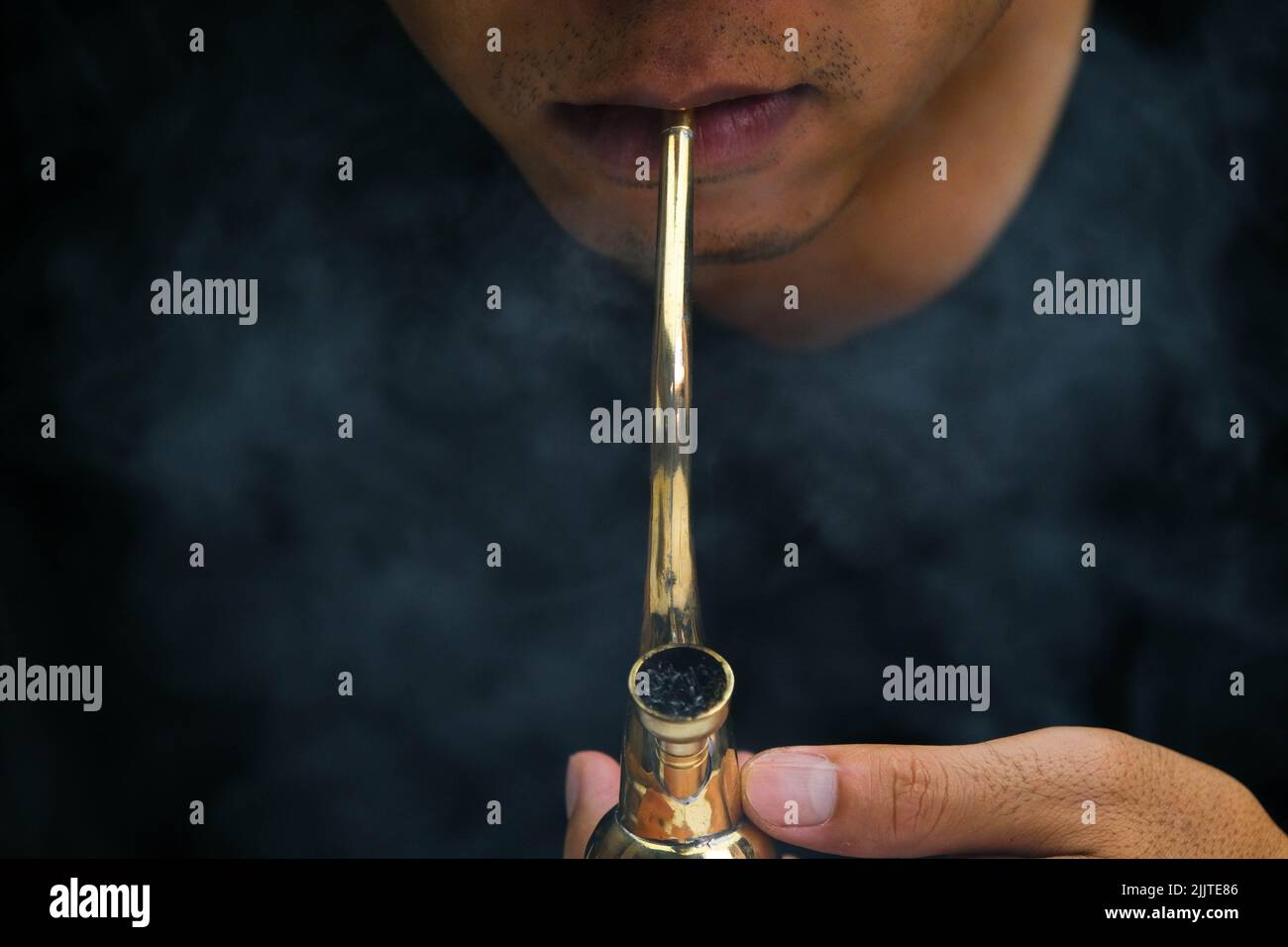 Ein asiatischer Mann raucht zu Hause aus einer Pfeife Marihuana. Studio-Shooting mit Model, das den Rauchtopf mit einer Pfeife vor dunklem Hintergrund simuliert. Legalisierung von Cannabis Stockfoto