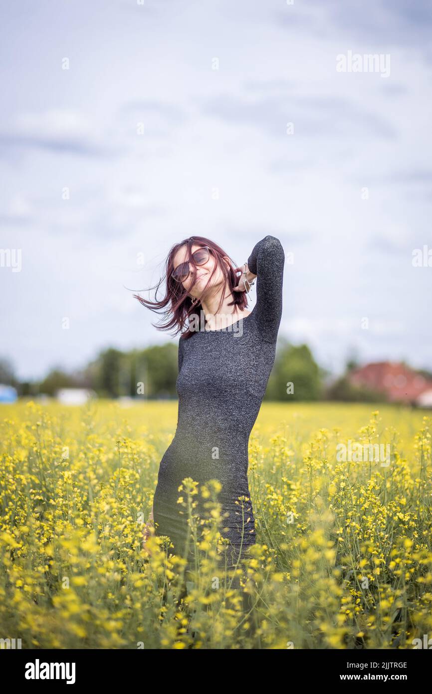 Eine schöne junge kaukasische Frau, die vor der Kamera posiert, während sie an einem sonnigen Tag zwischen blühenden gelben Blumen auf dem Feld steht Stockfoto