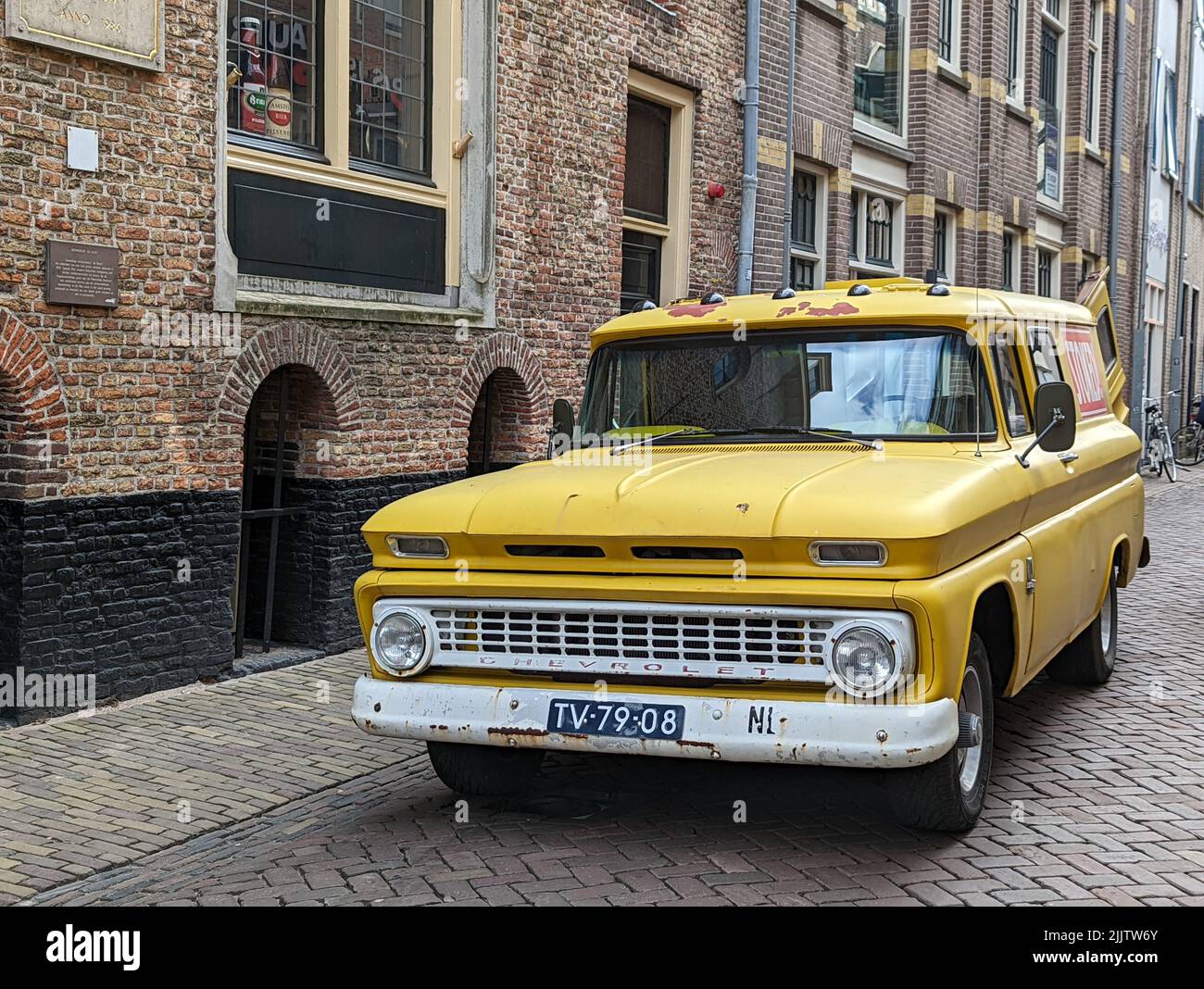 Eine szenische Aufnahme eines gelben Chevrolet Lieferwagens mit offener Heckklappe auf einer gepflasterten Straße mit traditionellen alten Backsteinhäusern Stockfoto