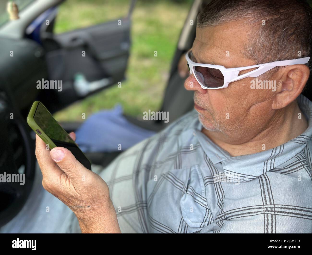 Schönes Porträt des ukrainischen Senior auf dem Fahrersitz, der im Sommer Nachrichten vom Handy-Bildschirm liest Stockfoto