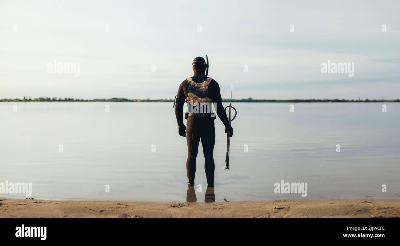 Rückansicht eines Mannes, der im Meerwasser steht und einen Neoprenanzug trägt und eine Speerpistole hält. Abenteuerlicher junger Mann, der sich bereit für das Speerfischen unter dem Wasser gemacht hat Stockfoto