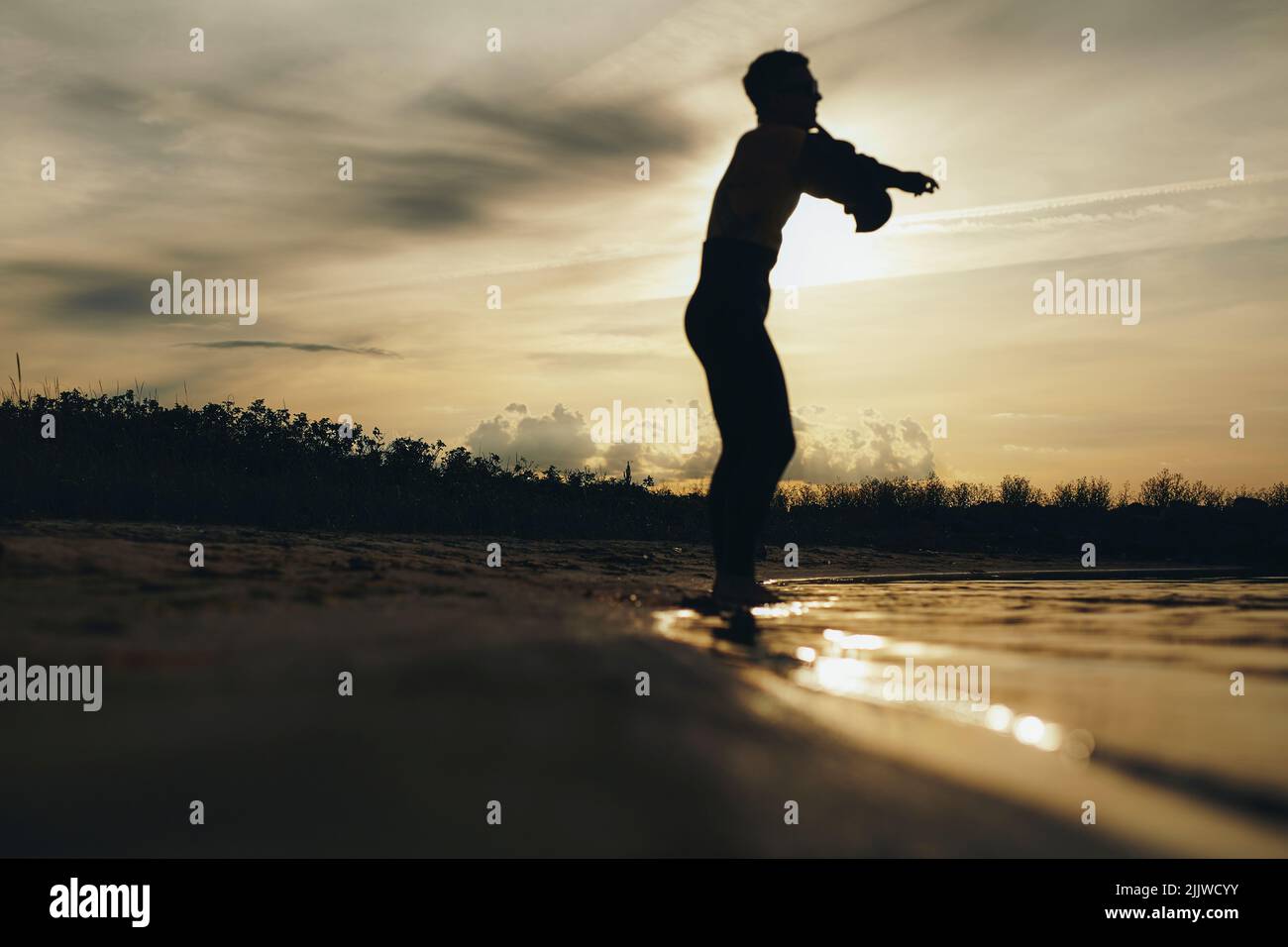 Silhouette eines männlichen Tauchers, der einen Neoprenanzug ansetzt, während er bei Sonnenuntergang am Strand steht. Abenteuerlicher junger Mann, der sich vorbereitet, um durch t ins Wasser zu kommen Stockfoto
