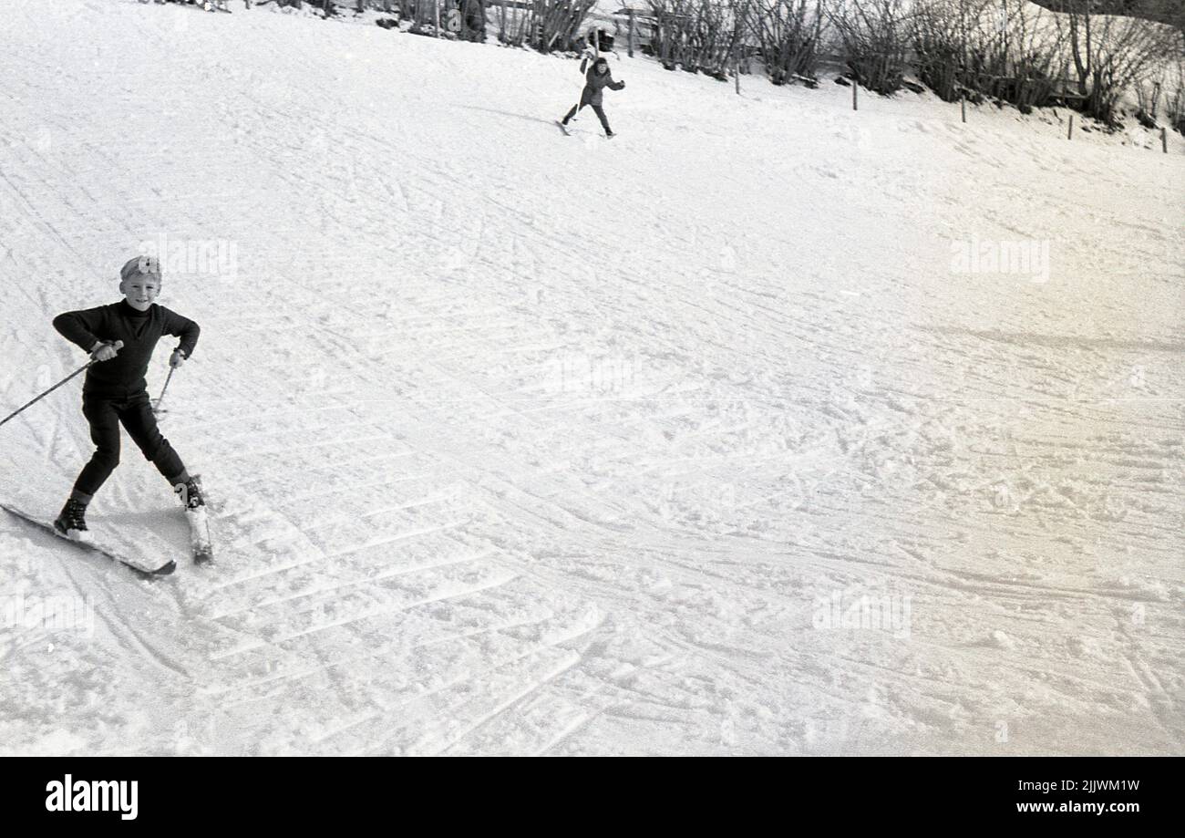 1966, historisch, winterlich und draußen auf einem Hügel, zwei kleine Kinder, die Ski fahren, wobei der Junge seine Skier zeigt, als er zum Stillstand kommt, England, Großbritannien. Stockfoto