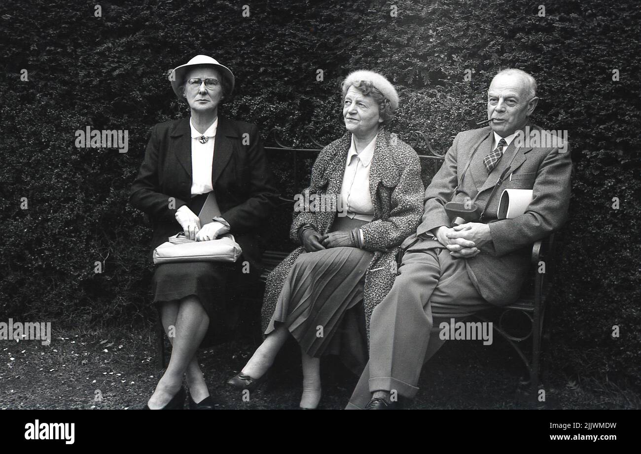 1960s, historisch, drei gut gekleidete ältere Menschen, die auf einer Parkbank sitzen, England, Großbritannien. Die Damen sind in eleganten Outfits, mit Handtaschen und Hüten, der Gentleman in Anzug und Krawatte, raucht eine Pfeife und hält eine Kamerahülle. Stockfoto