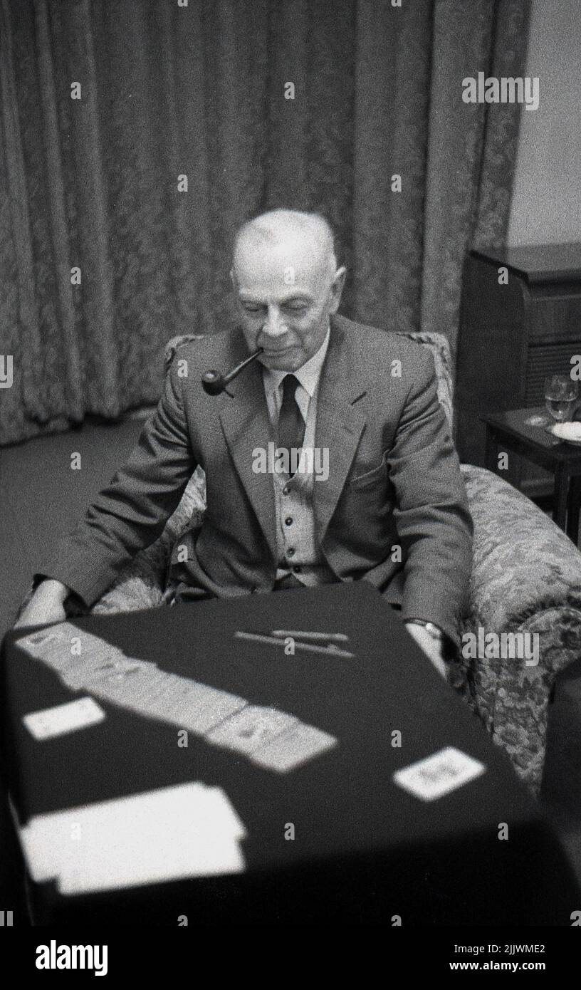 1960s, historisch, ein älterer Mann in Anzug und Krawatte und rauchig eine Pfeife, sitzend in einem Lounge-Stuhl an einem kleinen Tisch, der Karten spielt, England, Großbritannien. Stockfoto