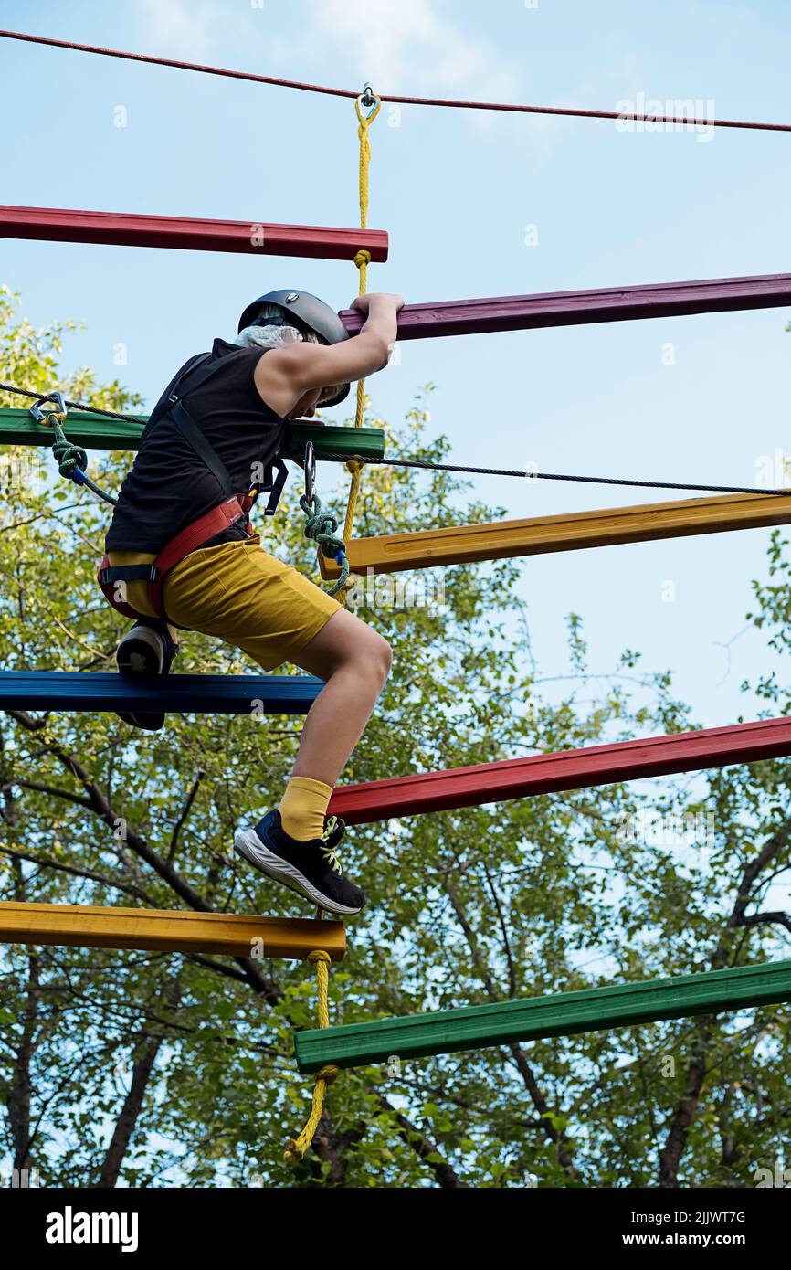 Kind im Sicherheitsgurt und Helm im Seilpark. Junge klettert mit Karabiner am Kabel auf Seilweg befestigt. Stockfoto