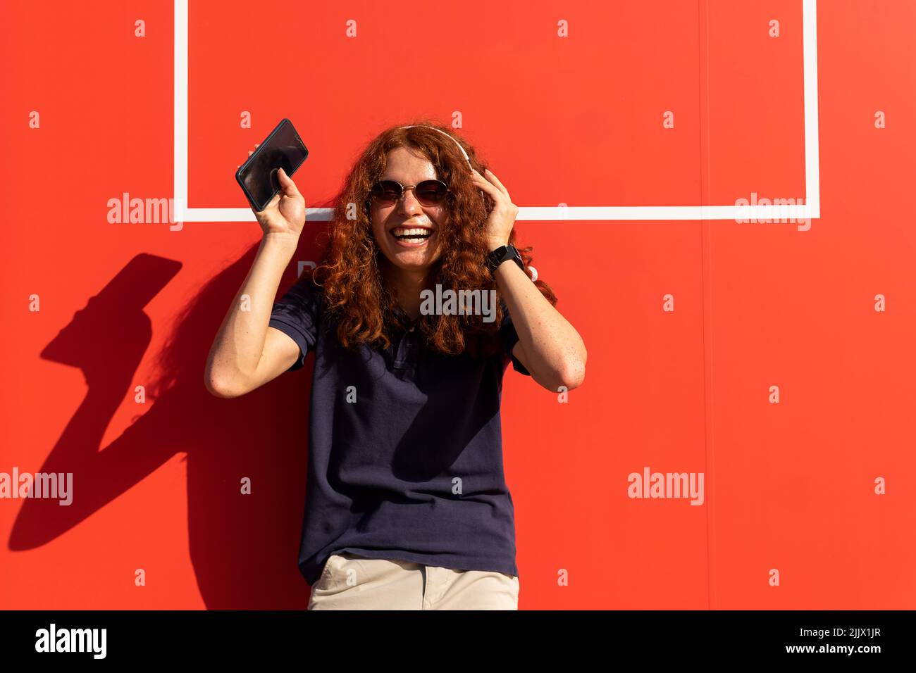 Lächelnde junge Frau mit Kopfhörern und Smartphone gegen die orangefarbene Wand Stockfoto