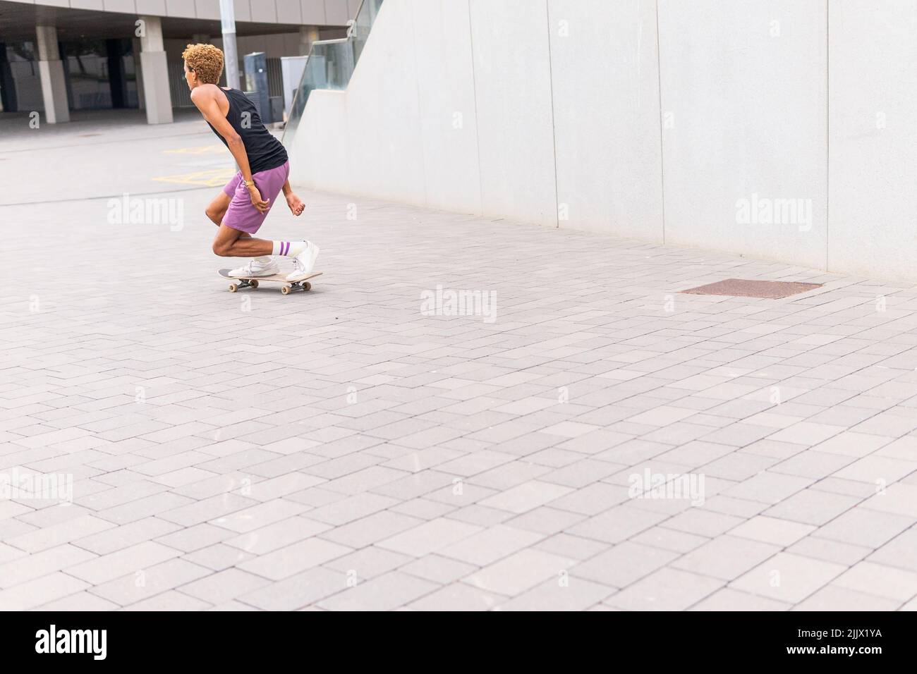 Ein ganzer Körper aktiver afroamerikanischer Männer, der während des Trainings auf der Straße der Stadt auf einem gepflasterten Gehweg in der Nähe des Gebäudes Skateboard reitet Stockfoto