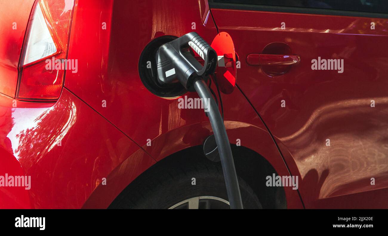 Gaspistole im Tank aus rotem Auto, das während des Betankens auf der Straße mit hellem Sonnenlicht in der Stadt auf der Tankstelle geparkt ist Stockfoto