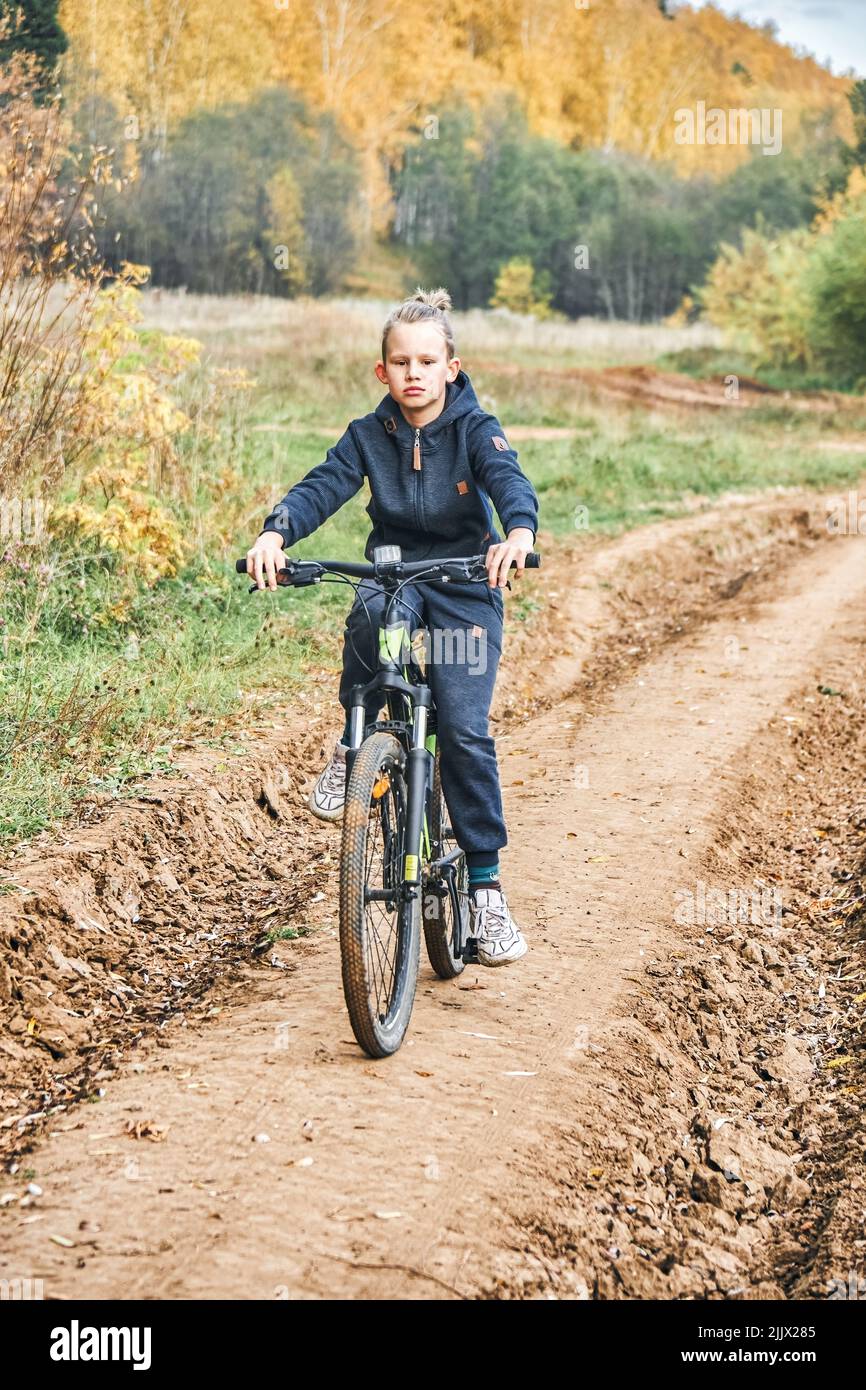 Der blonde Junge im lässigen Outfit fährt auf der Landstraße an Bäumen mit vergilbten Blättern vorbei auf dem Fahrrad. Schuljunge genießt sportliche Aktivitäten am Herbsttag Stockfoto