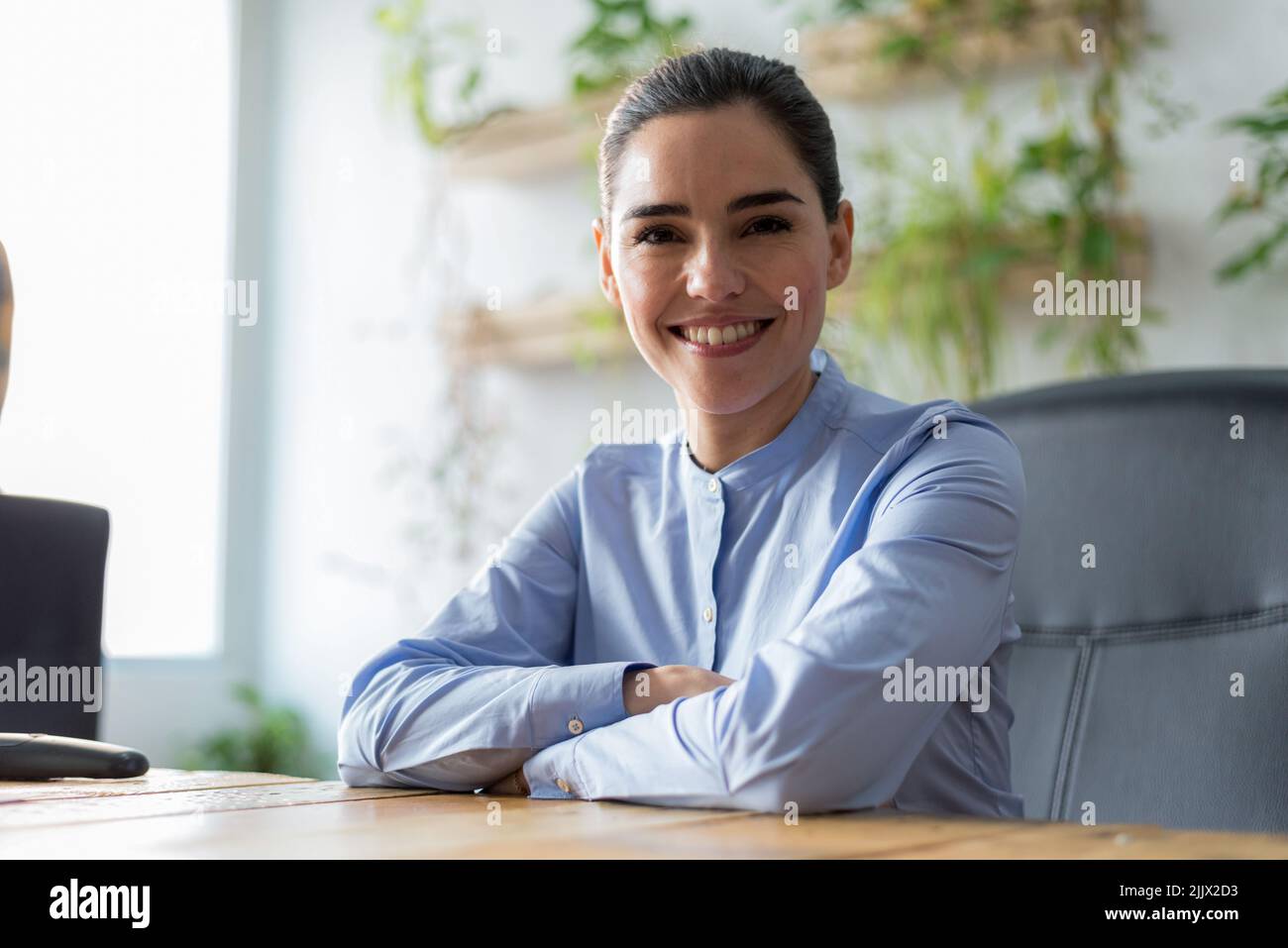 Fröhliche Managerin aus Kaukasien in weißer Bluse mit lockigen Haaren lächelnd und Blick auf die Kamera, während sie hinter dem Tisch im sonnendurchfluteten Büro sitzt Stockfoto