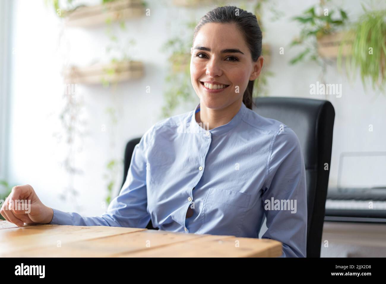 Fröhliche Managerin aus Kaukasien in weißer Bluse mit lockigen Haaren lächelnd und Blick auf die Kamera, während sie hinter dem Tisch im sonnendurchfluteten Büro sitzt Stockfoto