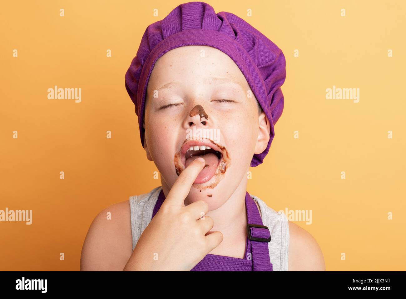 Inhalt chaotisch Junge mit geschlossenen Augen in lila Koch Uniform lecken Finger mit süßer Schokolade vor gelbem Hintergrund in Licht Studio Stockfoto